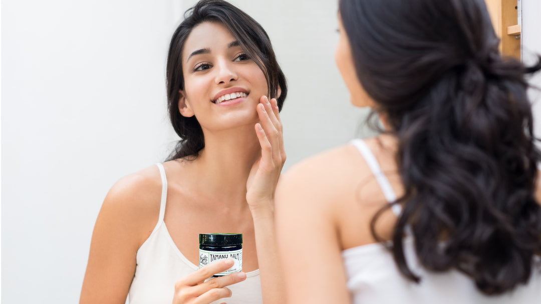 woman with long black hair looking at herself in the mirror holding a jar of erin's faces tamanu balm barrier salve to use for slugging