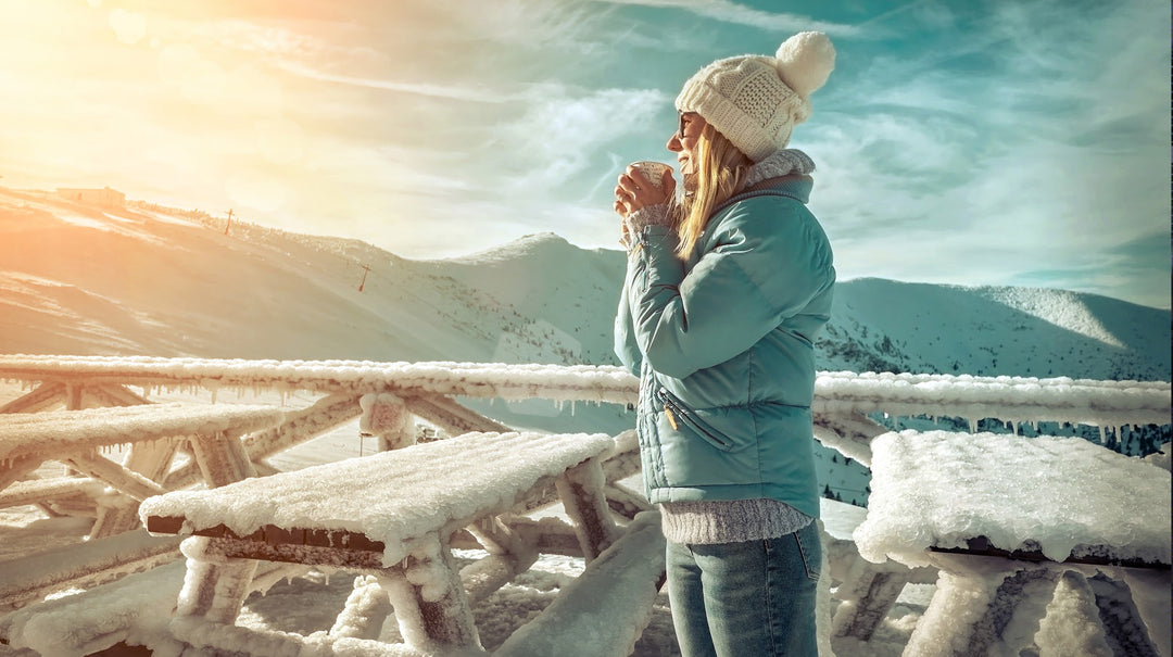 winter clad white woman sipping coffee surrounded by snow covered picnic benches overlooking sunrise and snow covered mountain