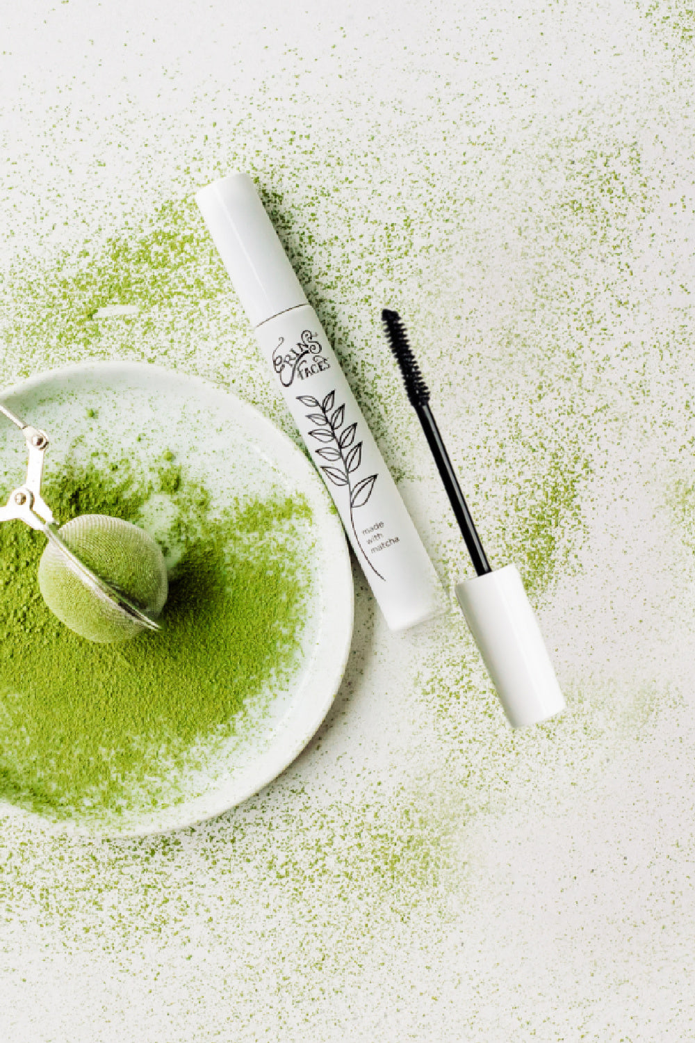 green tea, matcha, is powdered on a white surface with a white plate and tea sifter alongside white tubes of matcha mascara from erin's faces