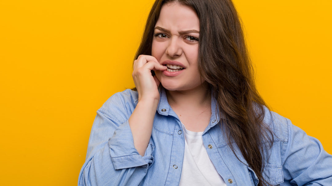white woman with long brown hair in denim shirt with yellow background chewing her nail nervously