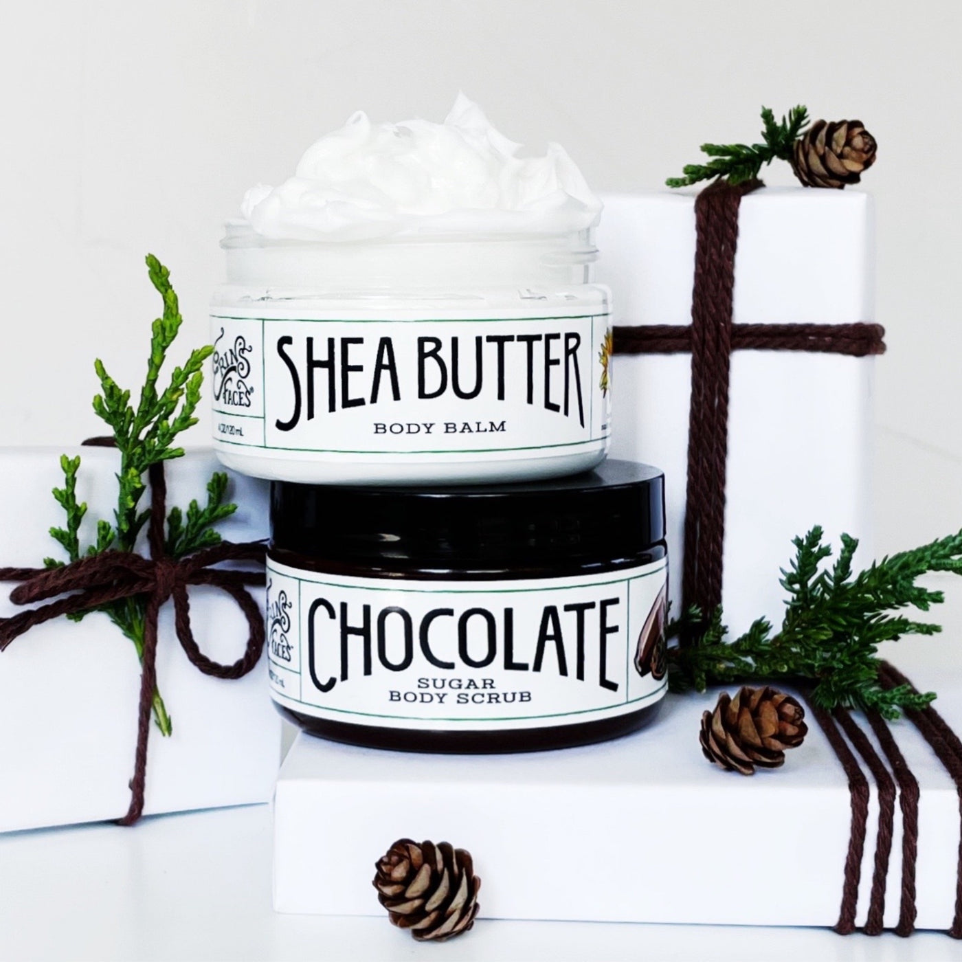 erin's faces shea butter body balm and chocolate sugar body scrub - the body balm is whipped so you can see it - surrounded by white paper packages tied up with brown string, bits of cedar and tiny pine cones