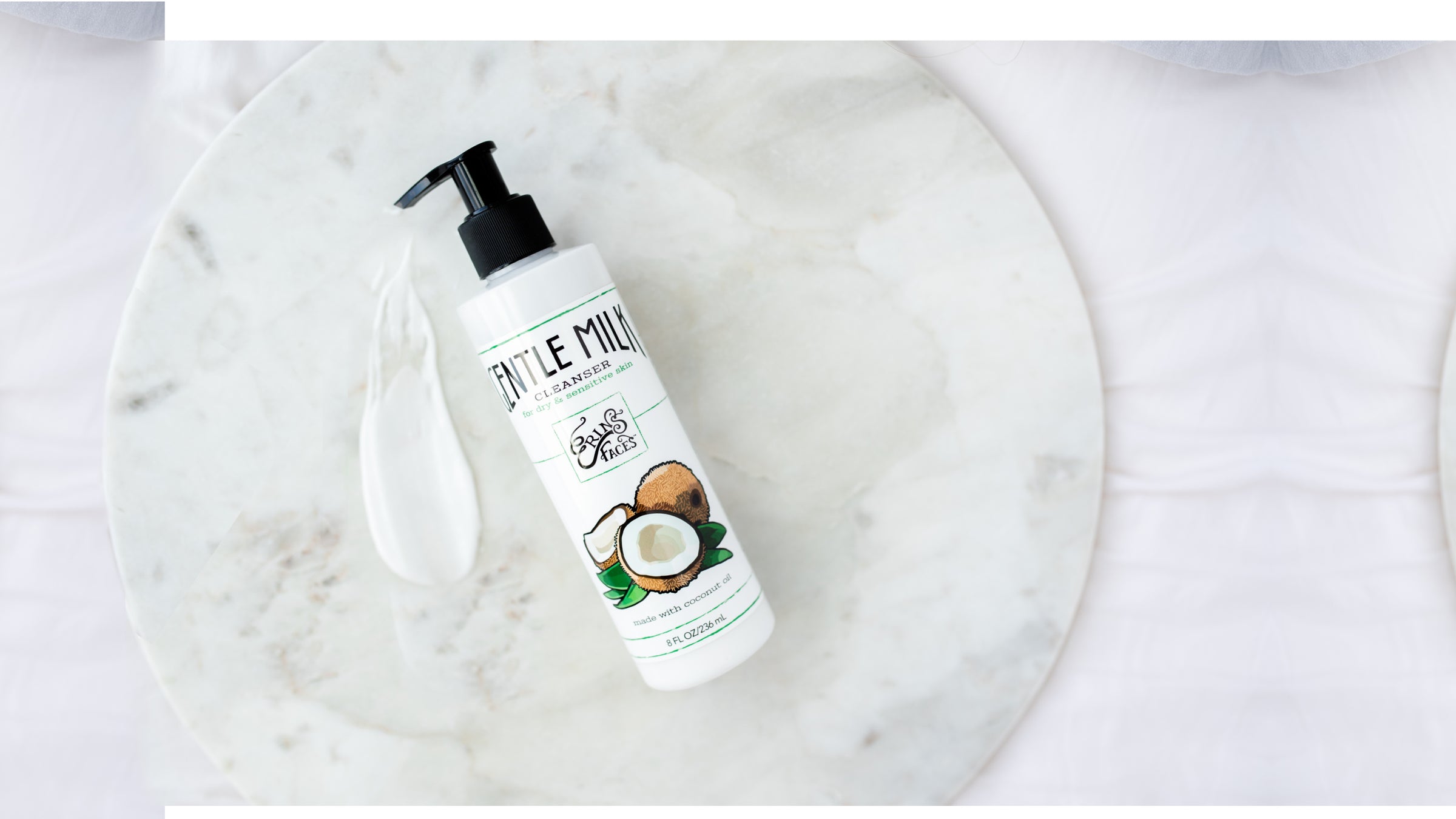 erin's faces gentle milk cleanser - bottle is white with coconut illustration - on white marble circle with swipe of product which is also white