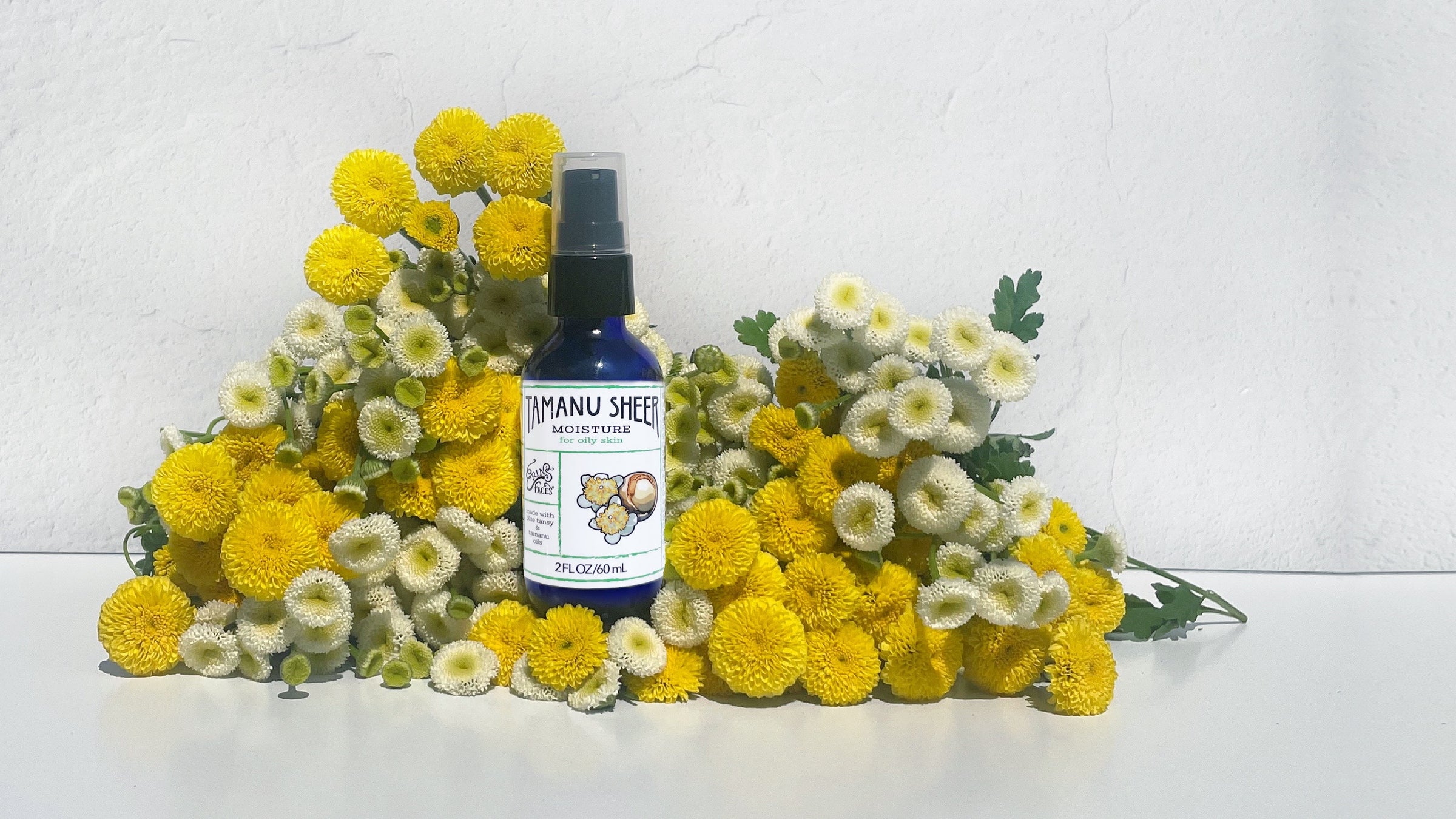 blue bottle of skincare (tamanu sheer moisture) on white background surrounded by tansy flowers that are yellow and white