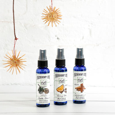 Blue plastic bottles of Erin's Faces Aromatherapy Sprays in Scotch Pine, Orange Clove and Cinnamon with straw ornaments against white bricks
