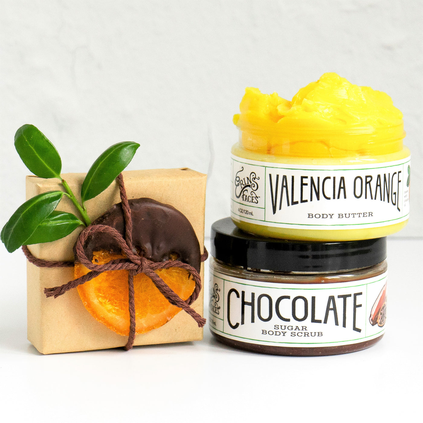 Valencia Orange Body Butter and Chocolate Sugar Body Scrub on white background next to brown paper gift wrapped box with brown string.  String holds a chocolate dipped dried orange slice and greenery