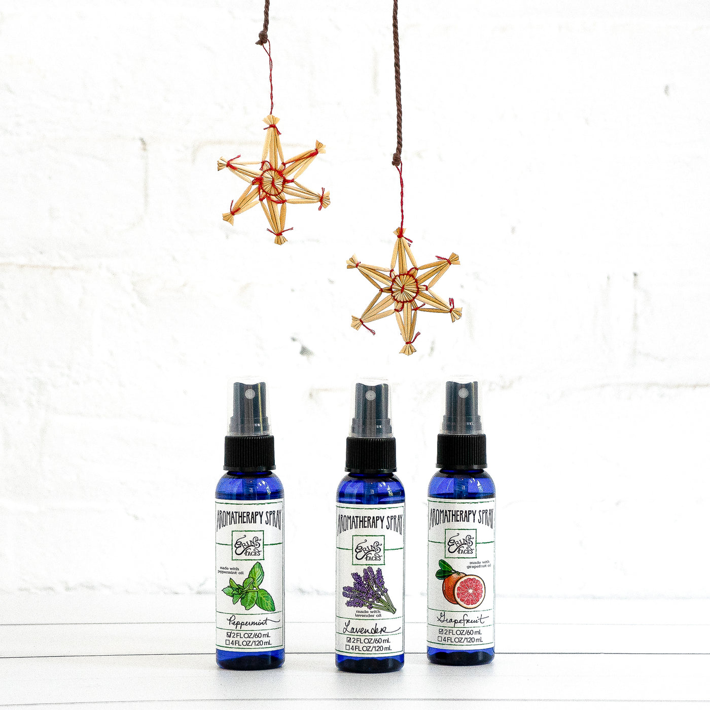 Blue plastic bottles of Erin's Faces Aromatherapy Sprays  of Peppermint, Lavender and Grapefruit with straw ornaments on a white brick background
