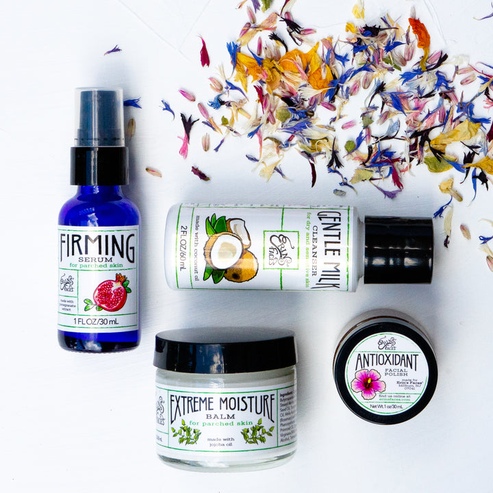 1oz blue glass bottle of firming serum, 2oz bottle of gentle milk cleanser, clear glass jar of extreme moisture balm, and 1oz mini jar of antioxidant facial polish on white background scattered with colorful flower petals