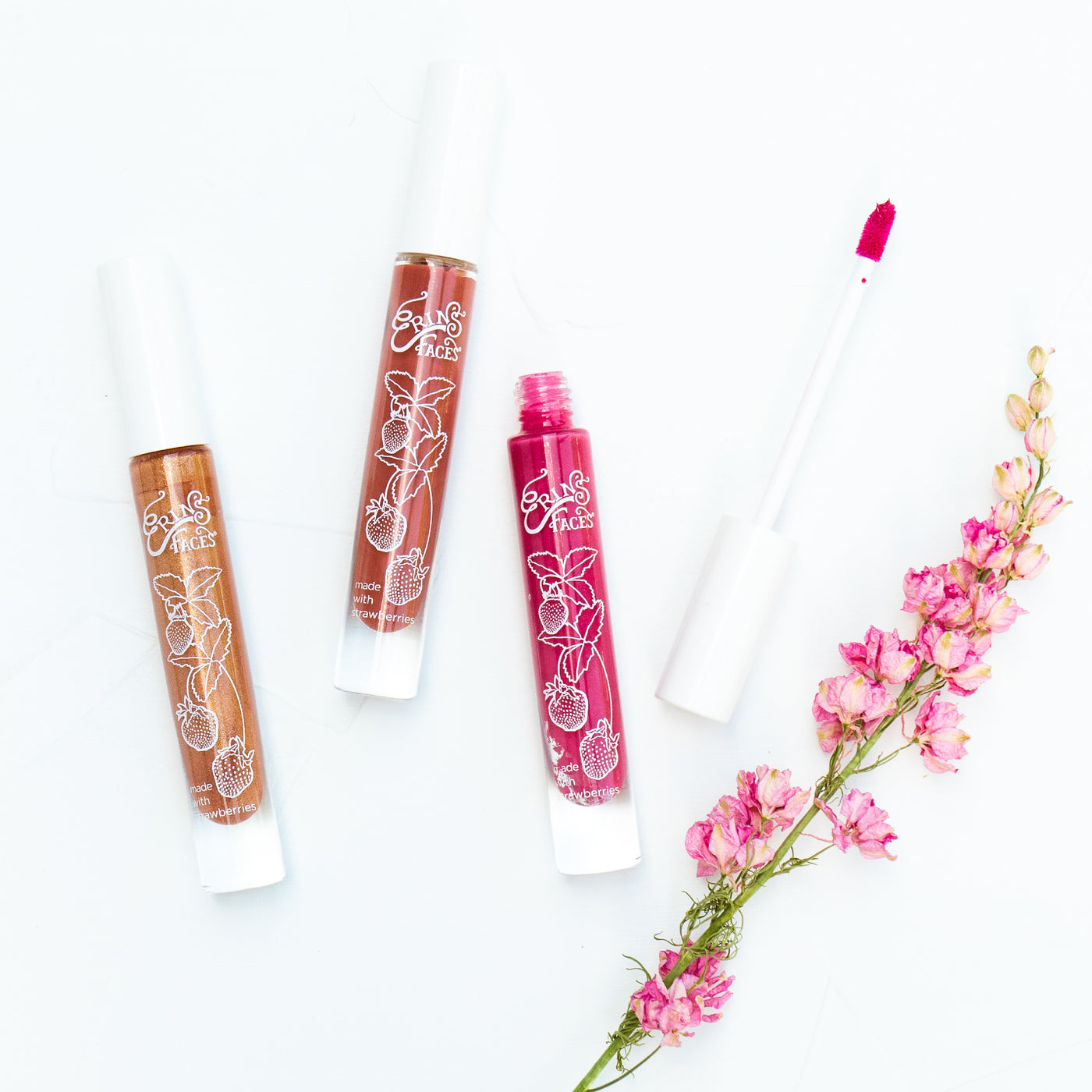 earliglow, sonata, eros fruit smoothie lip glosses from erin's faces with pink larkspur on white background