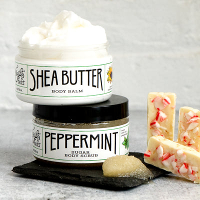 shea butter body balm jar on top of peppermint sugar body scrub with scrub sample next to bars of white chocolate with peppermint