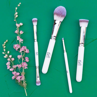 dried pink larkspur on a green background with white handled wooden brushes - double concealer, powder, angle and foundation